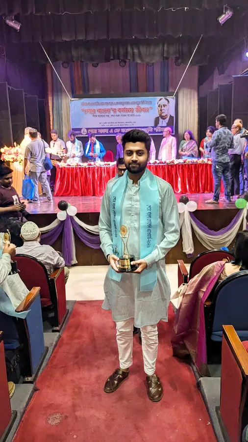 Mir Ahadul Islam standing in an auditorium in a pale green panjabi and award sash, holding the Sher-e-Bangla A.K. Fazlul Huq Golden Award trophy